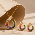 Gold crescent-shaped necklace and matching earrings with a row of rainbow-colored stones, displayed on a beige jewelry stand.
