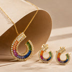 Gold crescent-shaped necklace and matching earrings with a row of rainbow-colored stones, displayed on a beige jewelry stand.