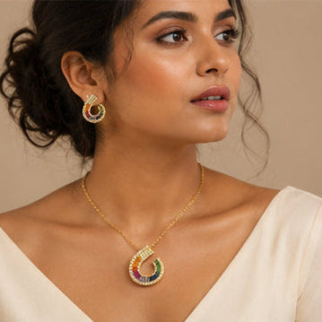 A woman wearing a gold crescent-shaped pendant necklace and matching drop earrings with colorful gemstone accents, styled with a cream-colored top against a warm beige background.