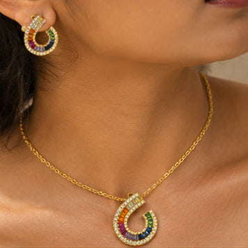 A close-up of a model wearing a gold horseshoe necklace and matching stud earring, both decorated with rainbow-colored gemstones and small clear crystals.