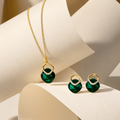 Gold jewelry set featuring a necklace and matching earrings with deep green heart-shaped crystals and small clear rhinestone accents, displayed on cream-colored curved stands.