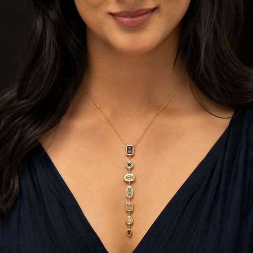 A close-up of a woman wearing a delicate gold necklace with a vertical drop design featuring multiple small gemstone charms. The necklace is shown against her dark navy blue V-neck top, with her dark hair framing her face.