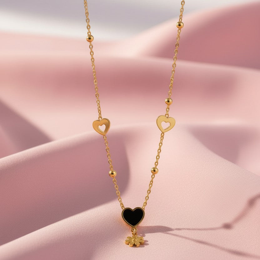 SVORA gold necklace with black enamel heart charm and daisy flower pendant, featuring hollow heart accents and gold beads.