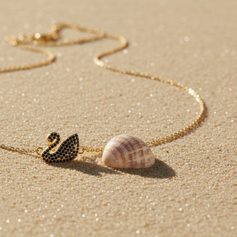 SVORA gold plated stainless steel necklace with a black swan pendant set with black CZ stones, on a sandy beach.