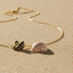 SVORA gold plated stainless steel necklace with a black swan pendant set with black CZ stones, on a sandy beach.