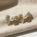 SVORA gold-tone bow stud earrings with pavé crystals and a central pearl, made of stainless steel.