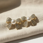 SVORA gold-tone bow stud earrings with pavé crystals and a central pearl, made of stainless steel.