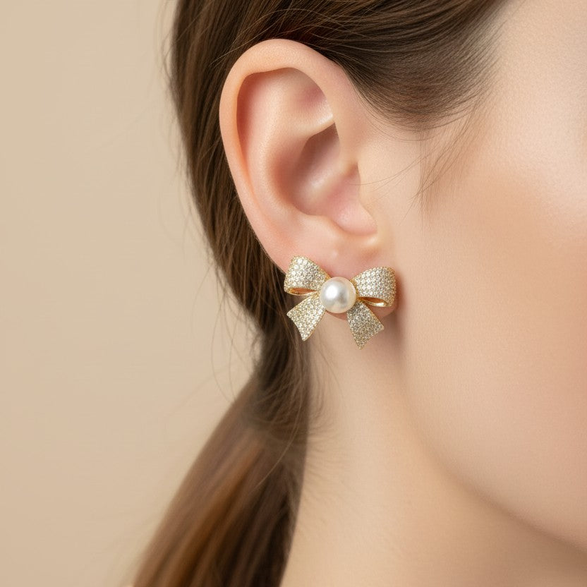 SVORA gold-tone bow stud earrings with pavé crystals and a central pearl, made of stainless steel.