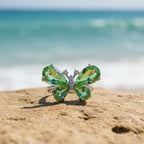 SVORA Ethereal Emerald Butterfly Ring with pear-cut green wings and pavé body on a beach.