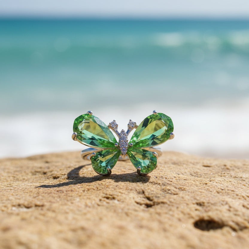 SVORA Ethereal Emerald Butterfly Ring with pear-cut green wings and pavé body on a beach.