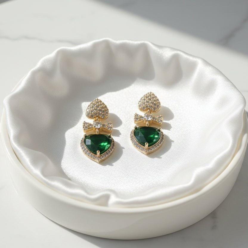 SVORA gold-tone drop earrings with pavé heart studs, bow connectors, and emerald crystal heart drops.