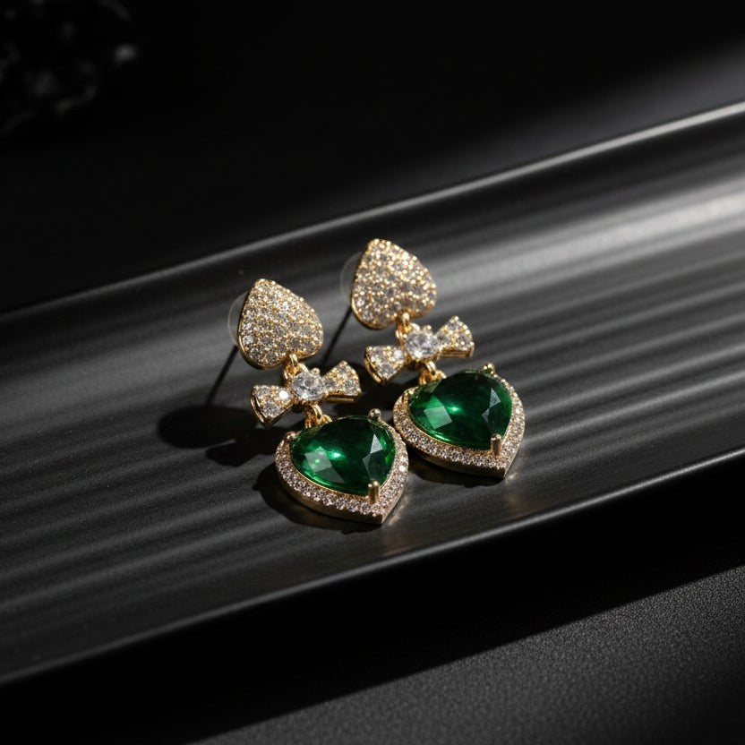 SVORA gold tone earrings with pavé heart studs, bow connectors, and emerald crystal heart drops.
