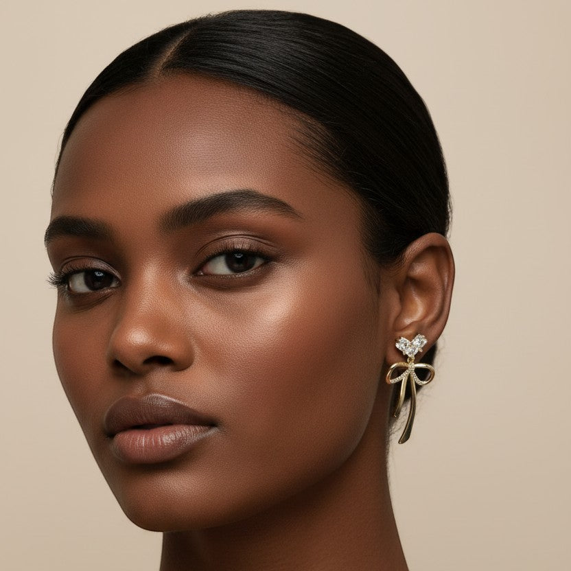 SVORA gold-tone drop earrings with crystal heart top and bow detail. Made with stainless steel and 18k gold plating.