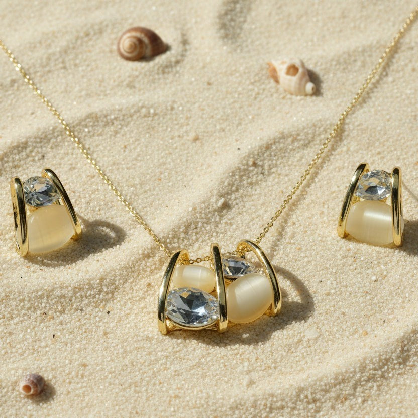 SVORA gold tone necklace and earrings set with clear cubic zirconia and cream cabochon stones in a caged design, resting on sand.