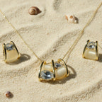SVORA gold tone necklace and earrings set with clear cubic zirconia and cream cabochon stones in a caged design, resting on sand.