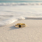 SVORA gold clover ring with black center and crystal white accents on a sandy beach with gentle waves.
