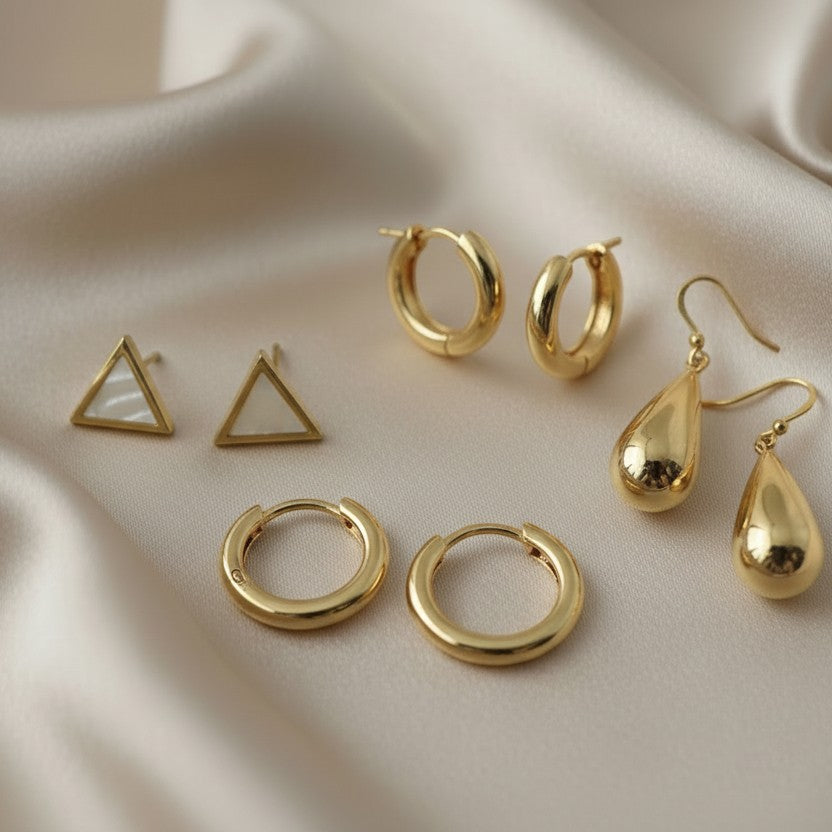 SVORA gold triangle studs with mother-of-pearl inlay, gold teardrop earrings, and gold hoop earrings on satin.