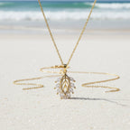 SVORA gold leaf cluster pendant necklace with marquise CZ stones on a delicate chain, set on a sandy beach.