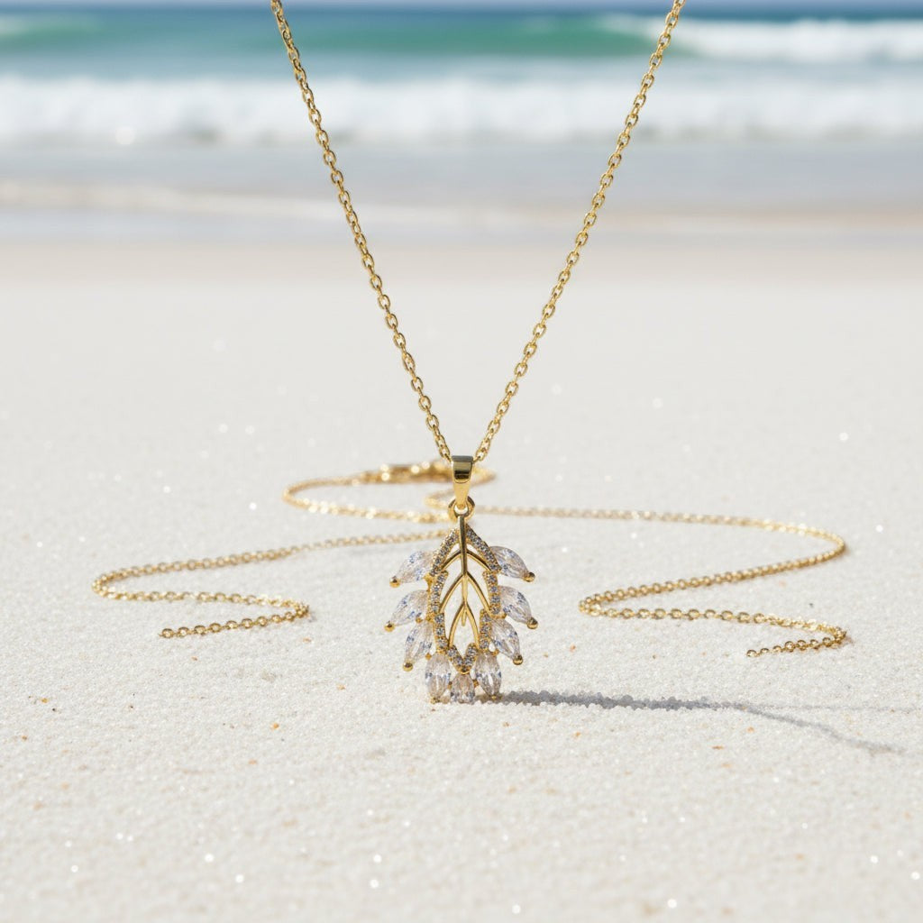 SVORA gold leaf cluster pendant necklace with marquise CZ stones on a delicate chain, set on a sandy beach.