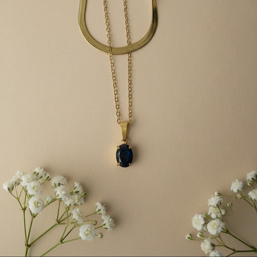 SVORA gold necklace with oval deep blue crystal pendant and snake chain, styled with baby's breath flowers.