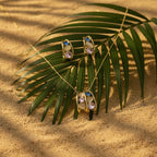 SVORA gold tone necklace and earrings set with pink crystals and blue accents, on a palm leaf over sand.