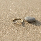 SVORA gold wishbone V-band ring with small diamonds on a sandy beach next to a smooth grey stone.