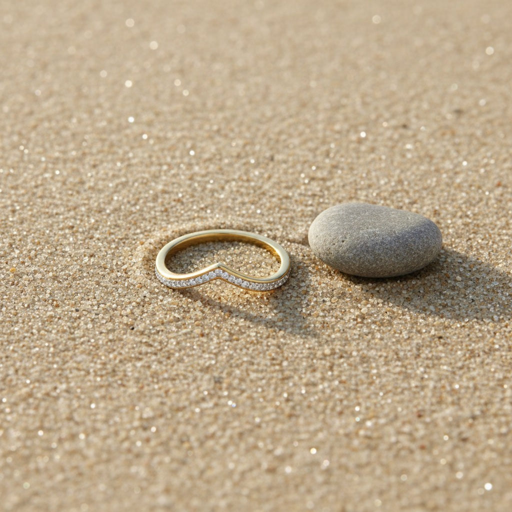 SVORA gold wishbone V-band ring with small diamonds on a sandy beach next to a smooth grey stone.