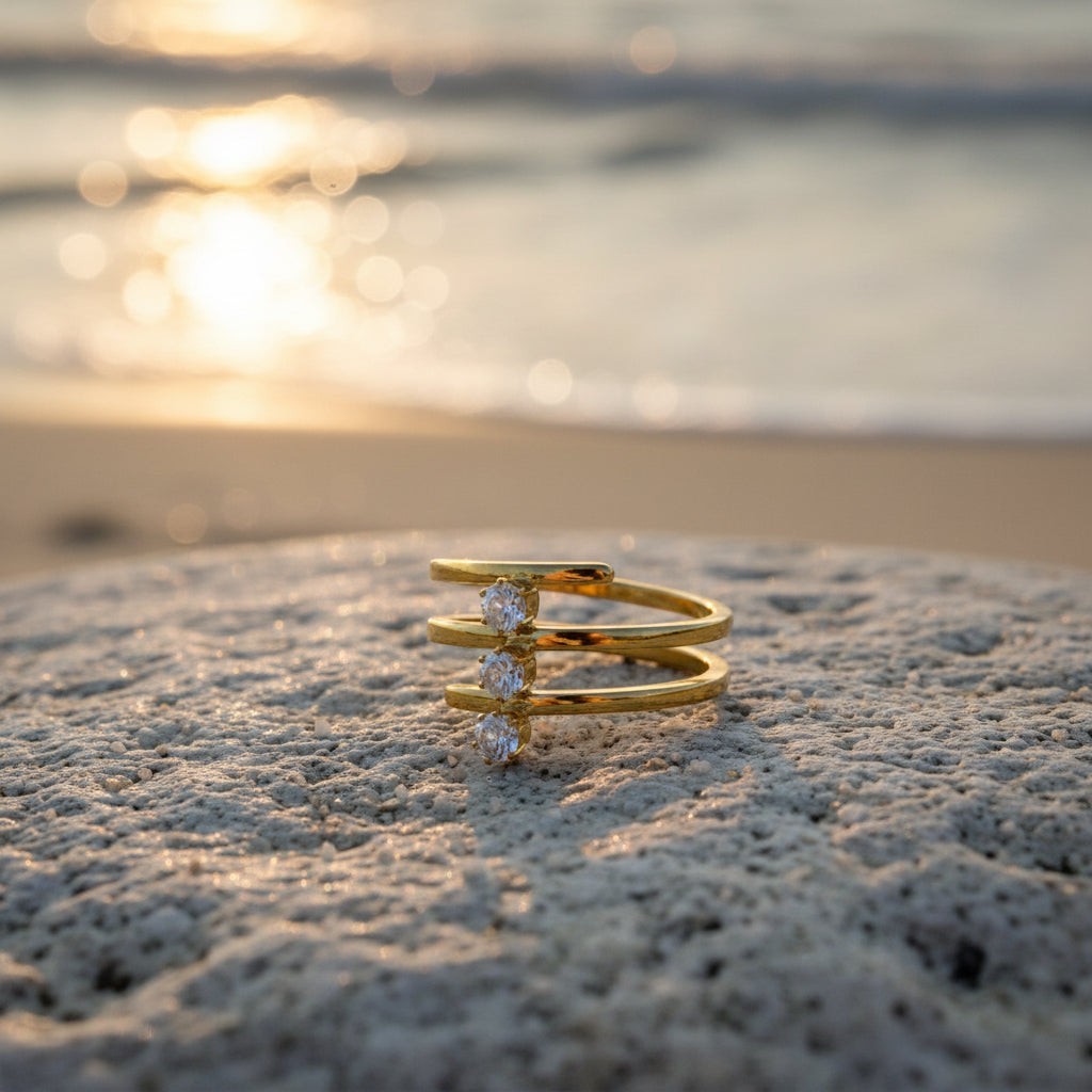 SVORA Lumiere Trilogy Spiral Ring in gold tone stainless steel with three cubic zirconia stones, set on a rock by the sea.