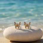SVORA mini gold butterfly stud earrings with cubic zirconia stones on a beach stone.