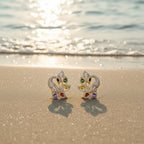 SVORA gold-tone peacock stud earrings with multicolour cubic zirconia stones on a sandy beach.