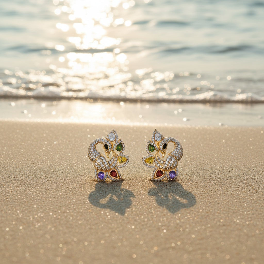 SVORA gold-tone peacock stud earrings with multicolour cubic zirconia stones on a sandy beach.