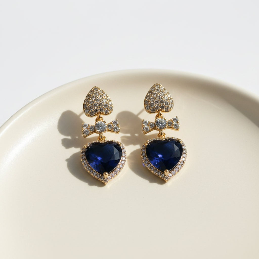 SVORA gold tone earrings with sapphire blue crystal heart drops and pavé bow connectors.